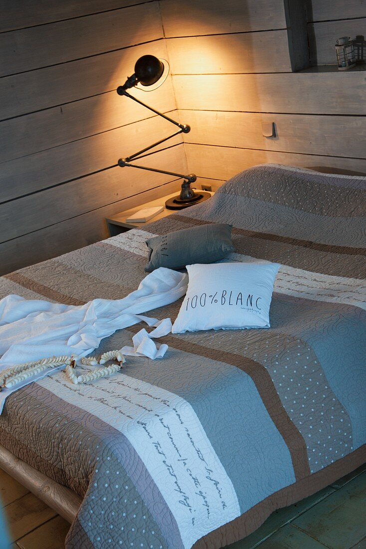 Doppelbett mit Tagesdecke in grau brauner Streifenoptik, im Hintergrund leuchtende Retro Tischlampe auf Nachttisch