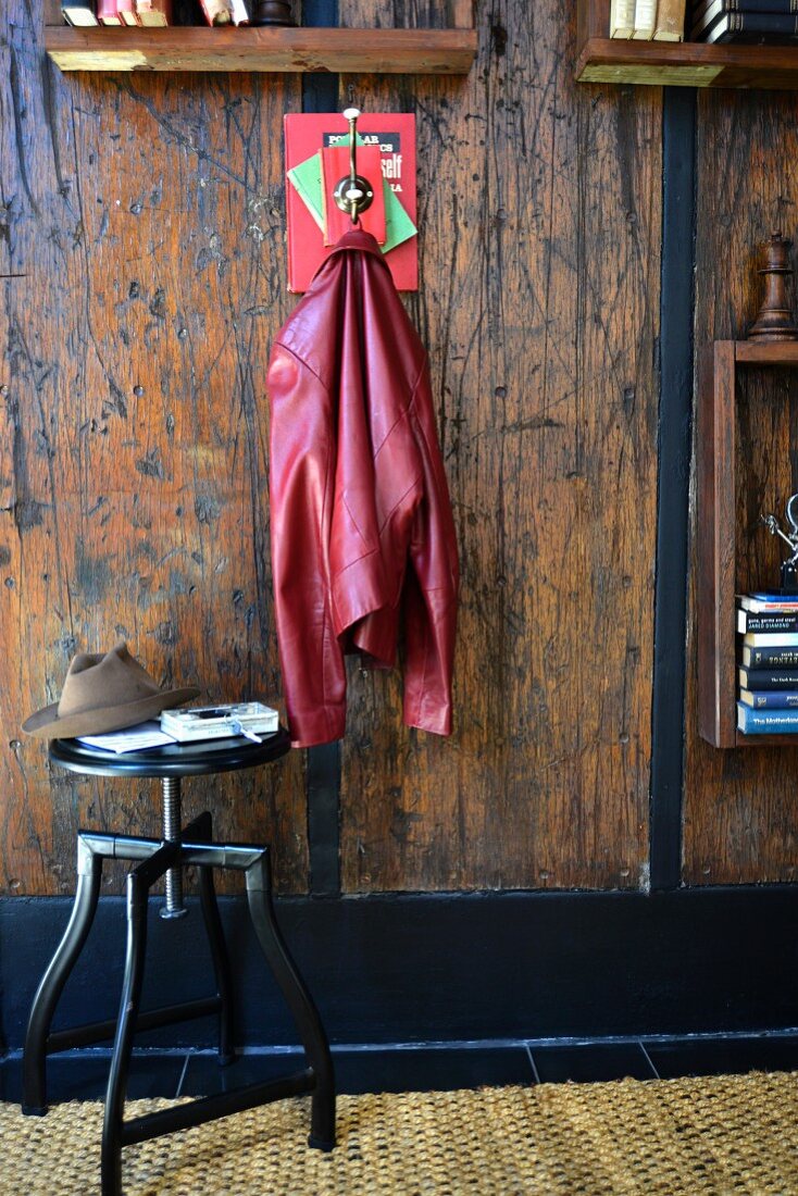 DIY-Garderobe aus Büchern und Kleiderhaken an rustikaler Holzwand mit Bücherregalen, dazu schwarzer Drehhocker mit Hut