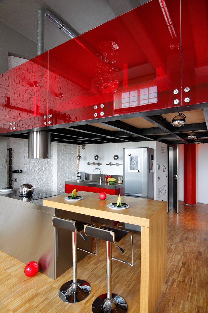 Wooden breakfast bar adjoining stainless steel island counter and bar stools below gallery with red glass balustrade in loft apartment