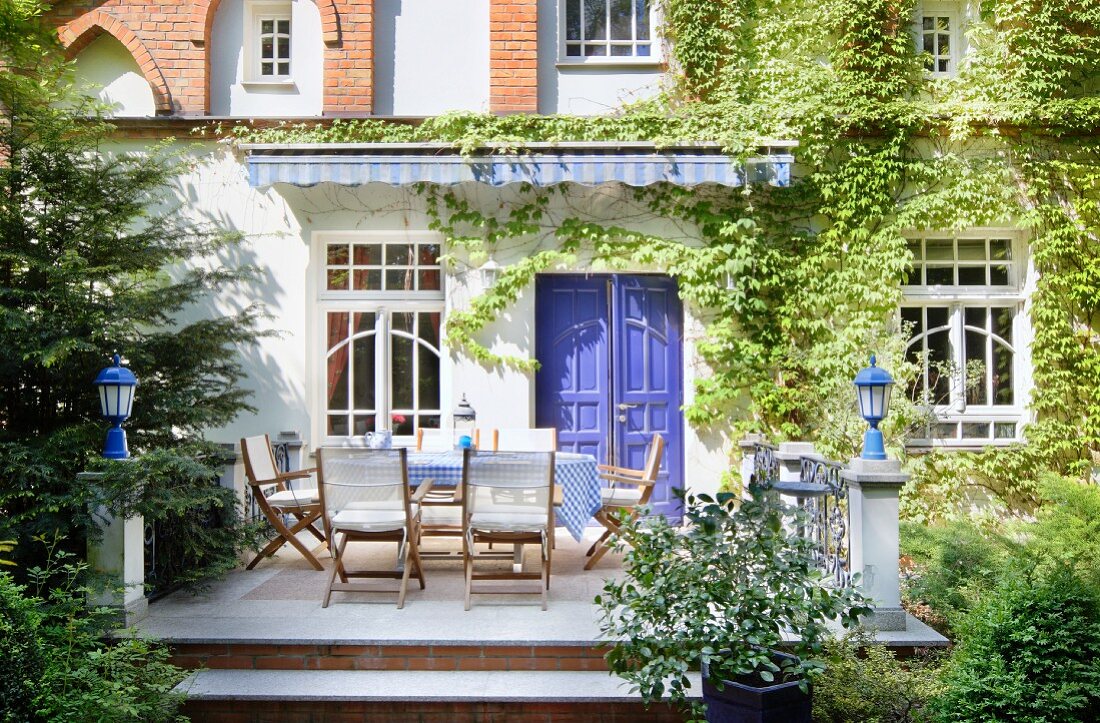 Blick vom Garten auf Terrasse mit Tisch und Stühlen, auf halbhohen Steinsäulen Laternen, im Hintergrund Wohnhaus mit berankter Fassade und blau lackierte Holztür