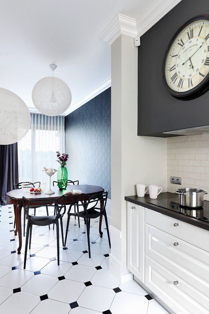 Open-plan kitchen with spherical lampshades above dining table, white kitchen counter in foreground, antique station clock and white and black tiled floor