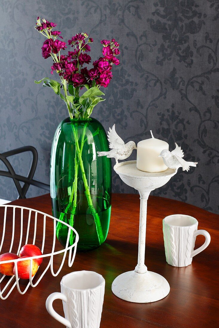 Stillleben mit grüner Glasvase, Retrodrahtkorb, weiße Porzellanbechern mit Strickmuster und Kerzenständer