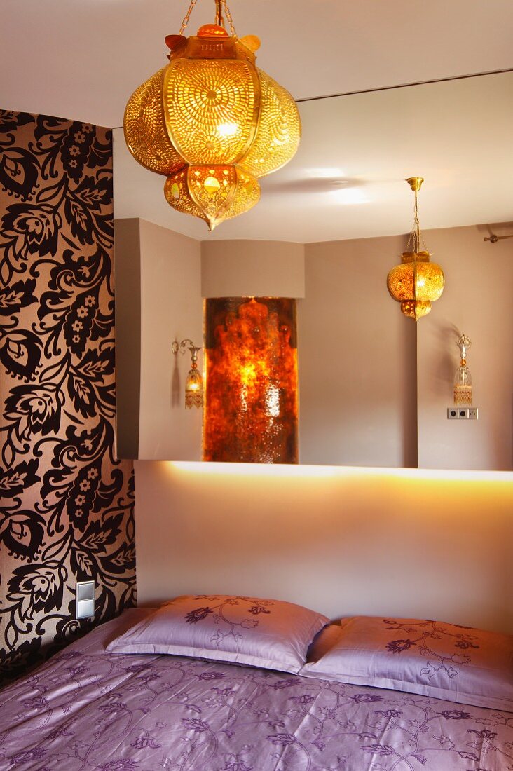 Spiegelwand über pastellviolett bezogenem Doppelbett, goldene orientalische Hängeleuchte und floral gemusterte Tapetenwand