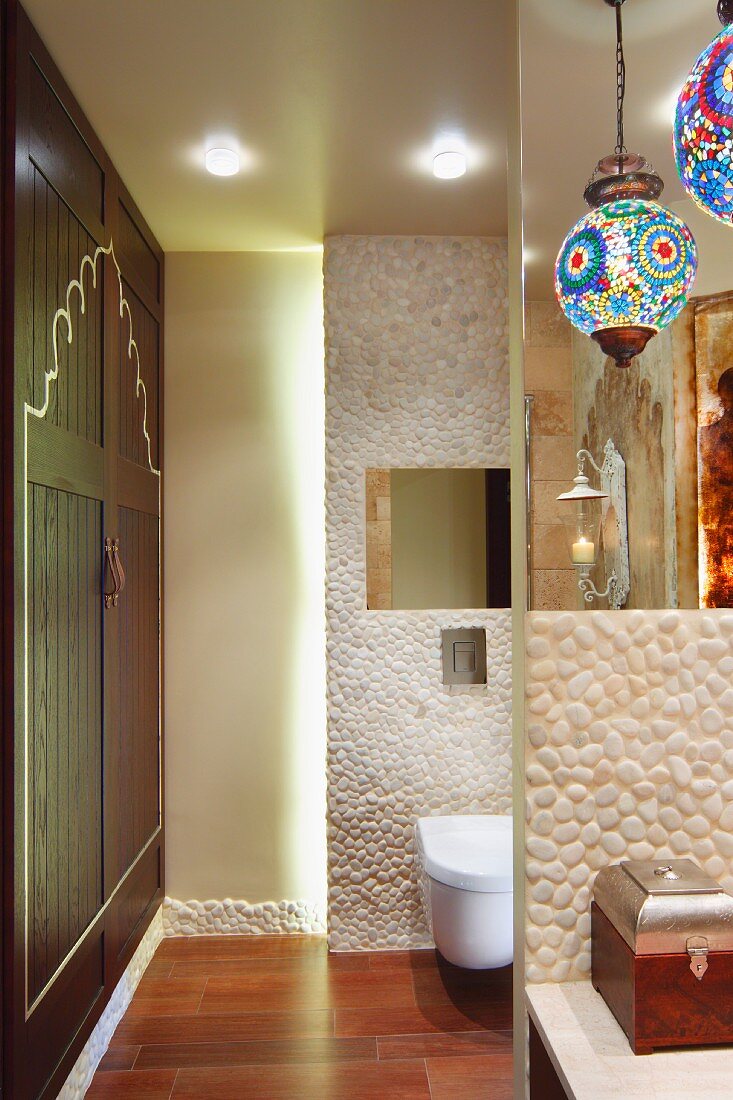 Bunte Kugelleuchten über Waschtisch mit Kiesel-Mosaikfliesen, dahinterliegendes WC-Abteil mit indirekter Beleuchtung; seitlich ein eingebauter Wandschrank