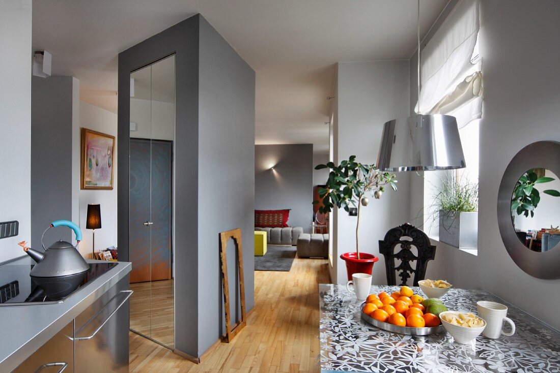Graue Küchenzeile mit Edelstahlfronten und Esstisch mit floralem Muster in der Glasplatte; Schrankskulptur als Raumteiler zum Wohnraum im Hintergrund