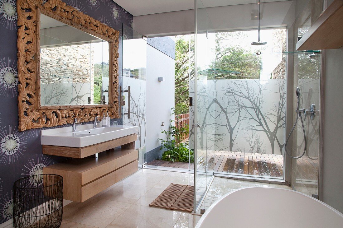 Verglaste, bodenebene Dusche, gegenüber Waschtisch mit Spiegel und Massivholzrahmen, mit geschnitzten Blättern, in modernem Bad, im Hintergrund Blick durch Glasfassade auf Terrasse