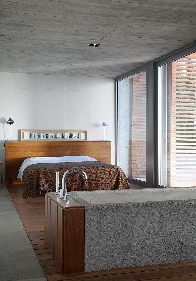 Badewanne aus Beton und Doppelbett mit brauner Tagesdecke im Schlafzimmer mit Betondecke