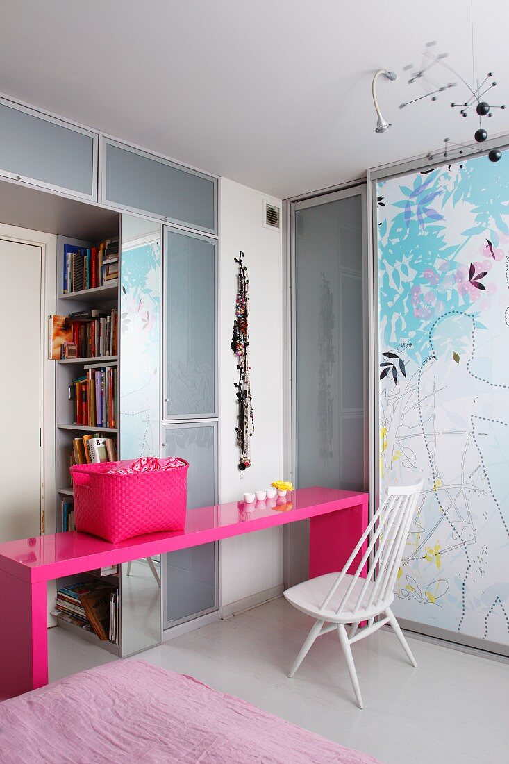 White wooden chair in front of modern, pink desk with colour-coordinated basket in corner of room with fitted cupboards