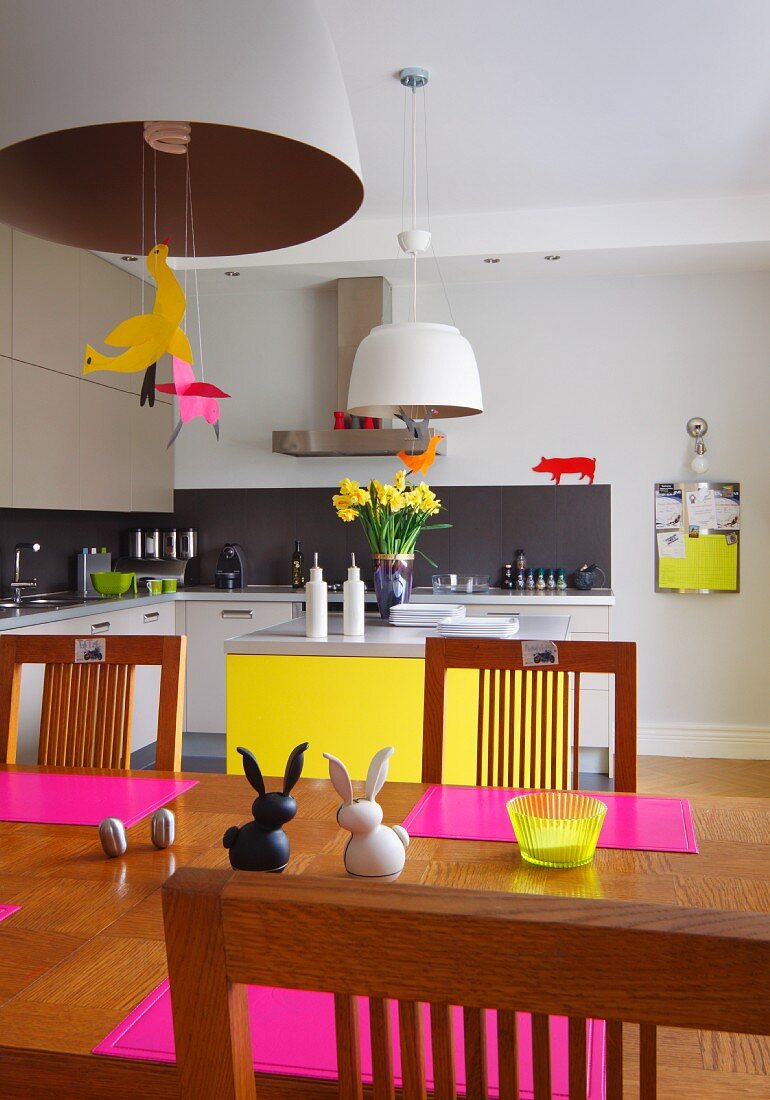 Bold pink and canary yellow accents in open-plan kitchen with dining area, Easter ornaments on dining table below pendant lamps and modern fitted cabinets in background