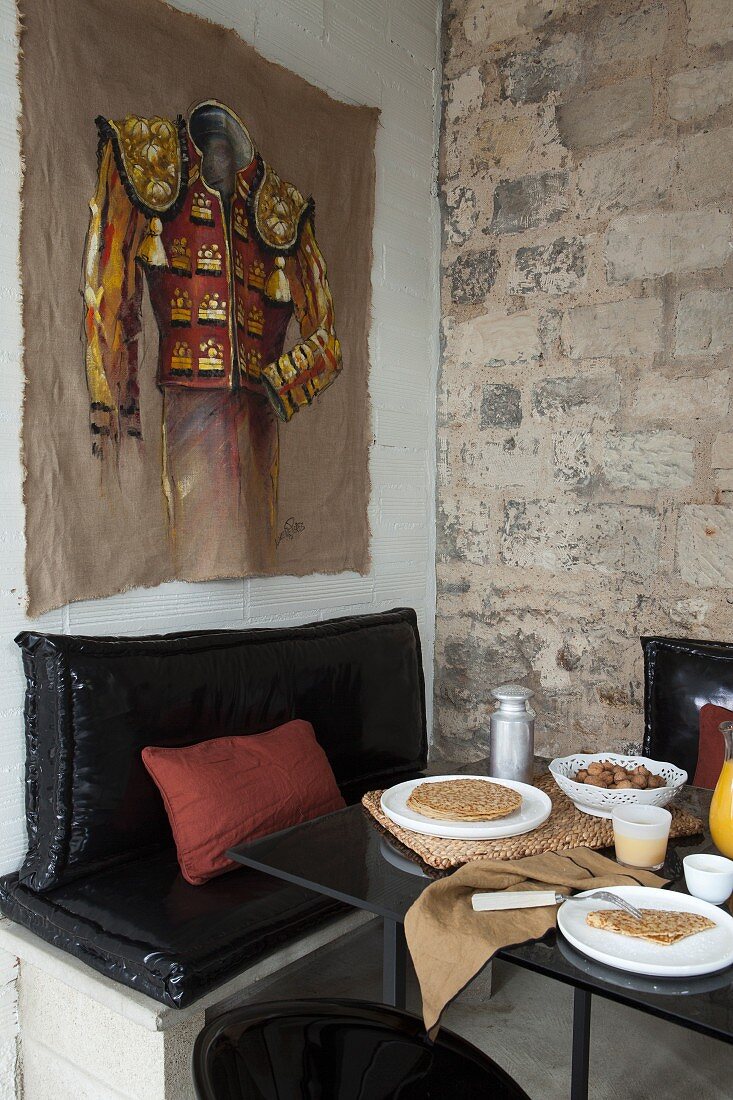 Essplatz in Vintage-Zimmerecke, Gemälde an Wand über schwarzem Sitzpolster auf gemauertem Unterbau und gedeckter Tisch