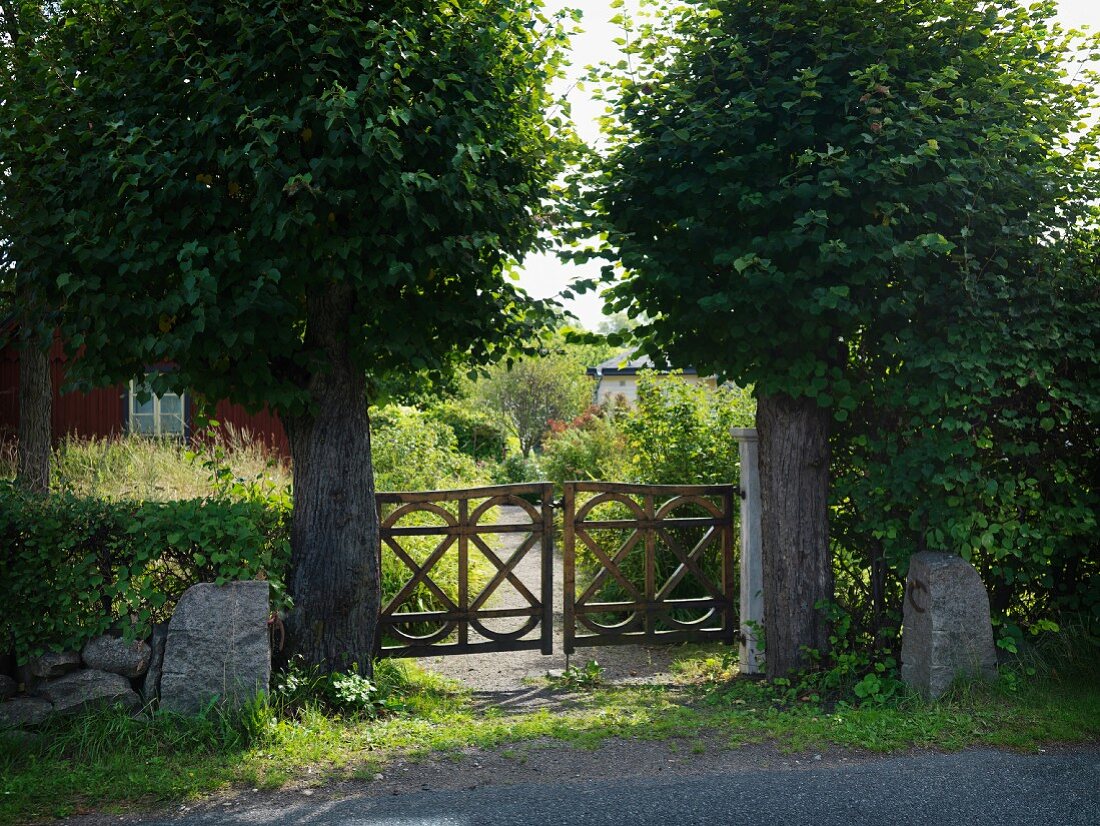 Blick von der Strasse auf Gartentor, in rustikaler Holzausführung, umgeben von Bäumen mit kugelförmiger Baumkrone
