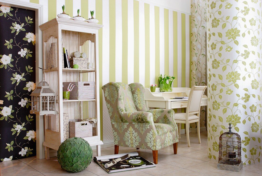 Interior in mixture of pretty patterns with white bureau, armchair, open-fronted shelving unit and walls covered in striped & floral wallpapers