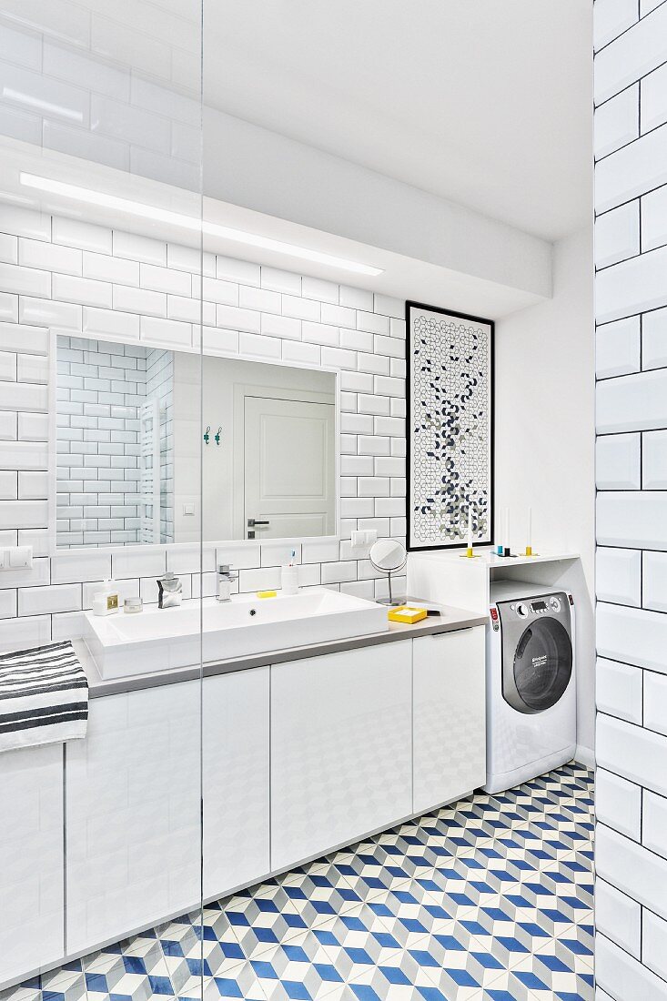 Blick in modernes weisses Badezimmer mit Wand- & Bodenfliesen, Waschtisch mit Spiegel, Lichtleiste & Unterschränken, im Hintergrund Waschmaschine in Überbauschrank