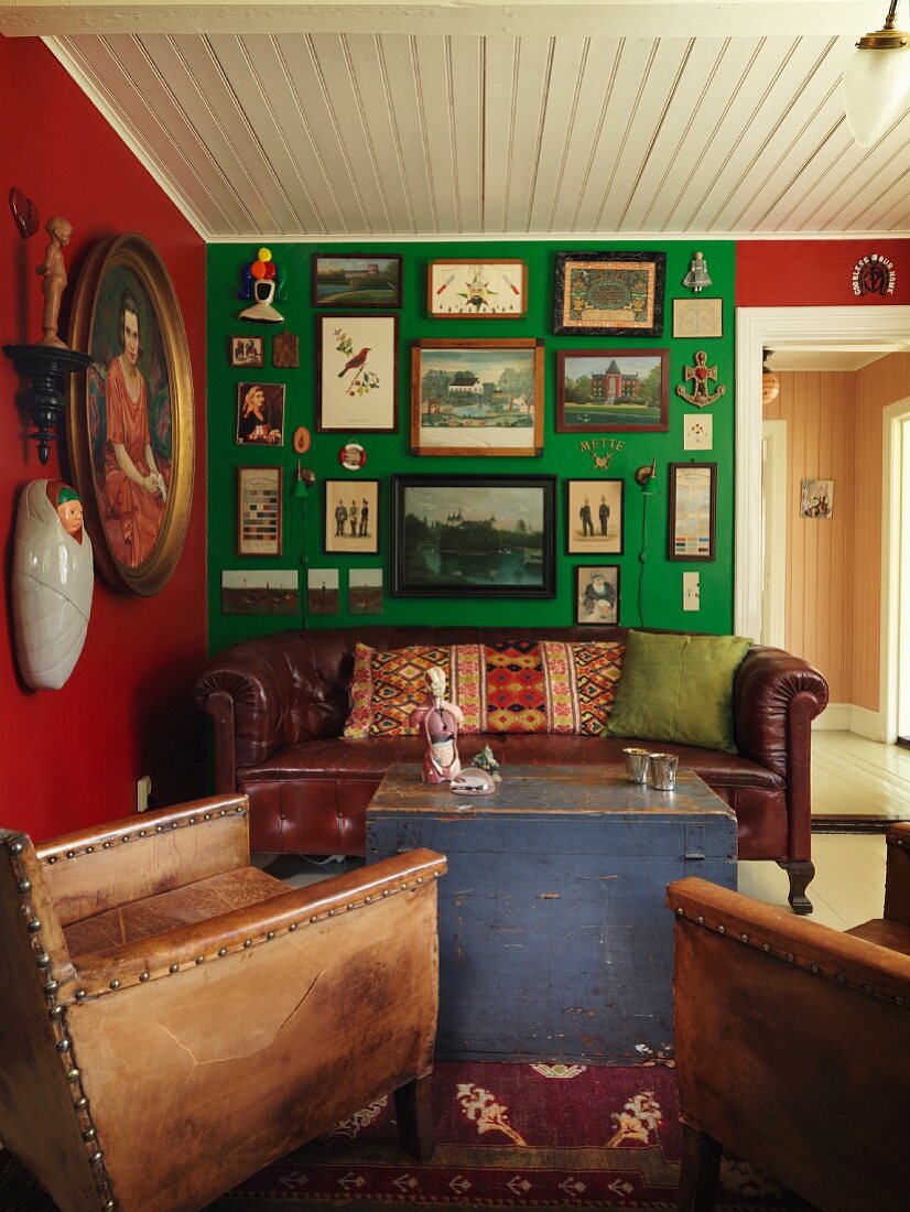 Vintage leather armchair and antique leather couch around wooden trunk used as coffee table in corner of living room; walls in rich colours decorated with gallery of pictures
