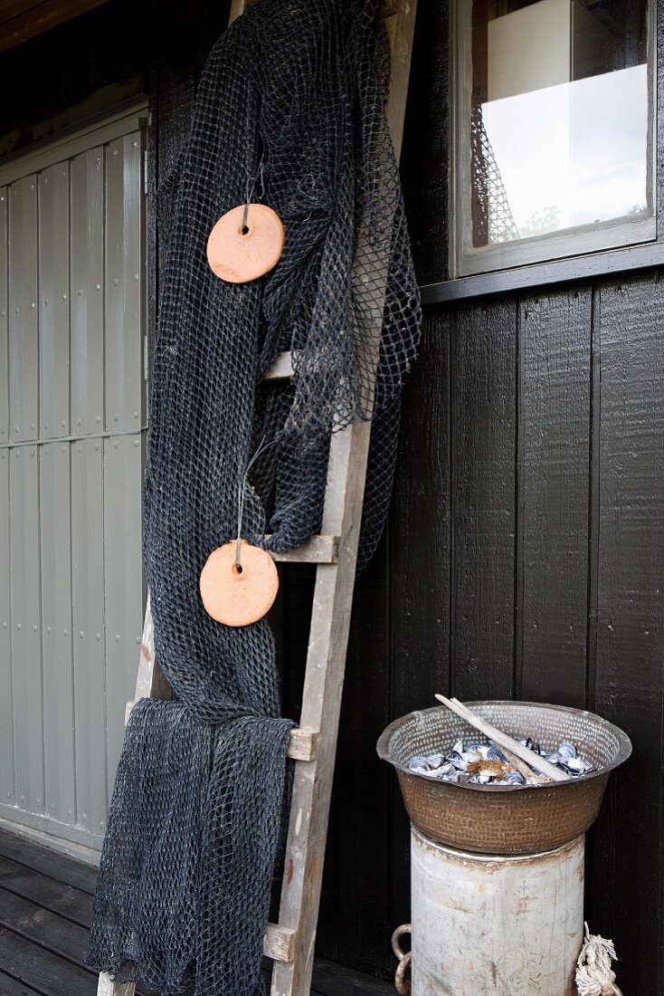 Fishing nets draped over ladder next to dish of seashells on cylindrical tub against house façade