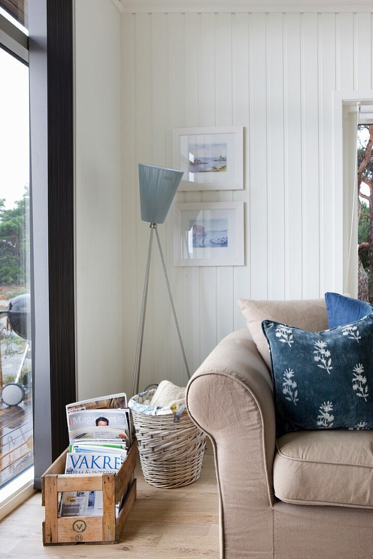 Scatter cushions on sofa, standard lamp and crate of magazines in corner of white, wood-clad living room