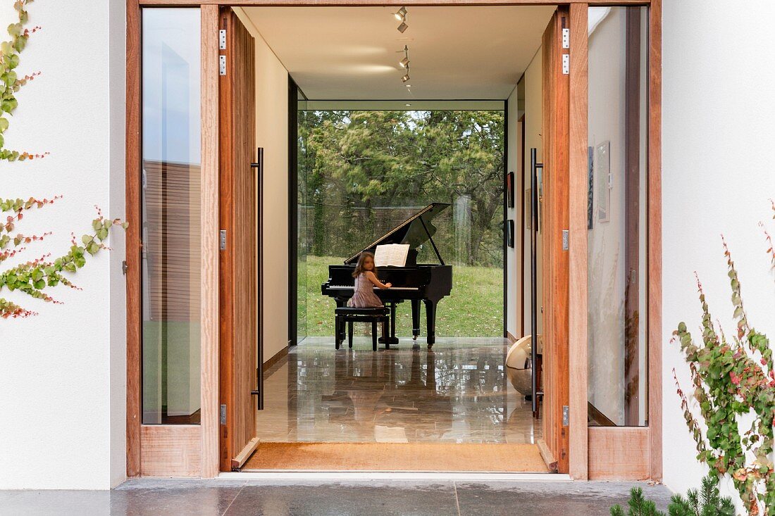 Blick durch offene Eingangstür in Foyer eines Wohnhauses, Mädchen an Klavierflügel mit Verglasung zum Garten