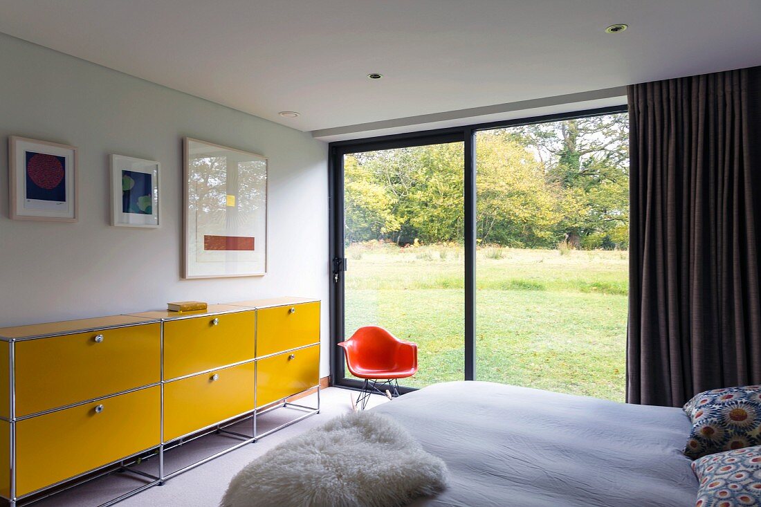 Schlafzimmer mit Stahlrohr-Sideboard in Gelb neben orangerotem Klassiker Schalenstuhl vor Glasfront mit Landschaftsgartenblick