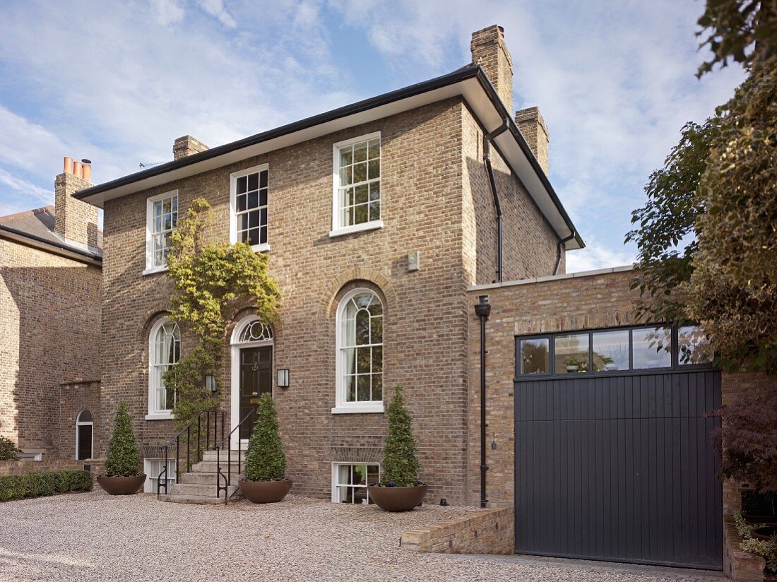 English house with brick facade, arched windows with white lattice frames on ground floor, narrow steps leading to front door and gravel front courtyard