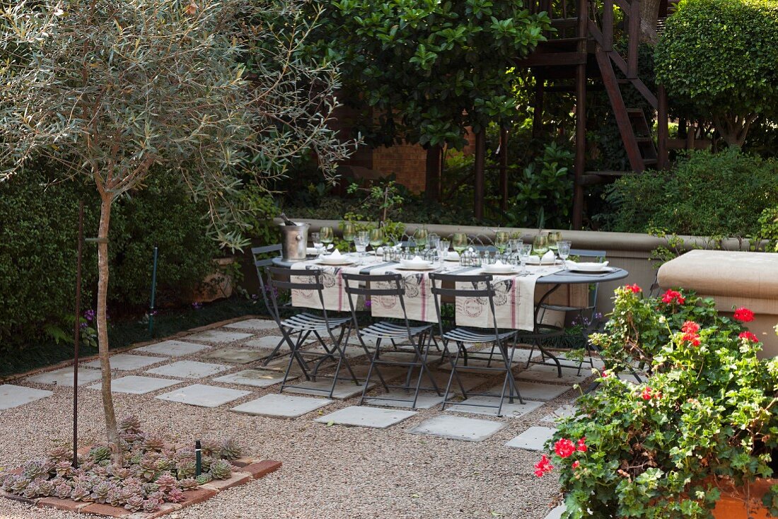 Gedeckter Tisch im Garten auf mit Kies & Steinplatten ausgelegtem Platz unter Bäumen