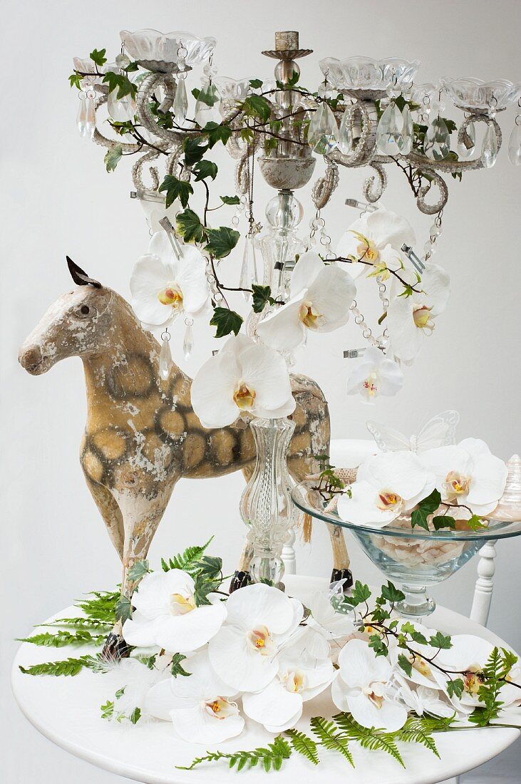 White orchids in a bowl and on a candle holder with glass decorations and an antique miniature horse behind it on a white table