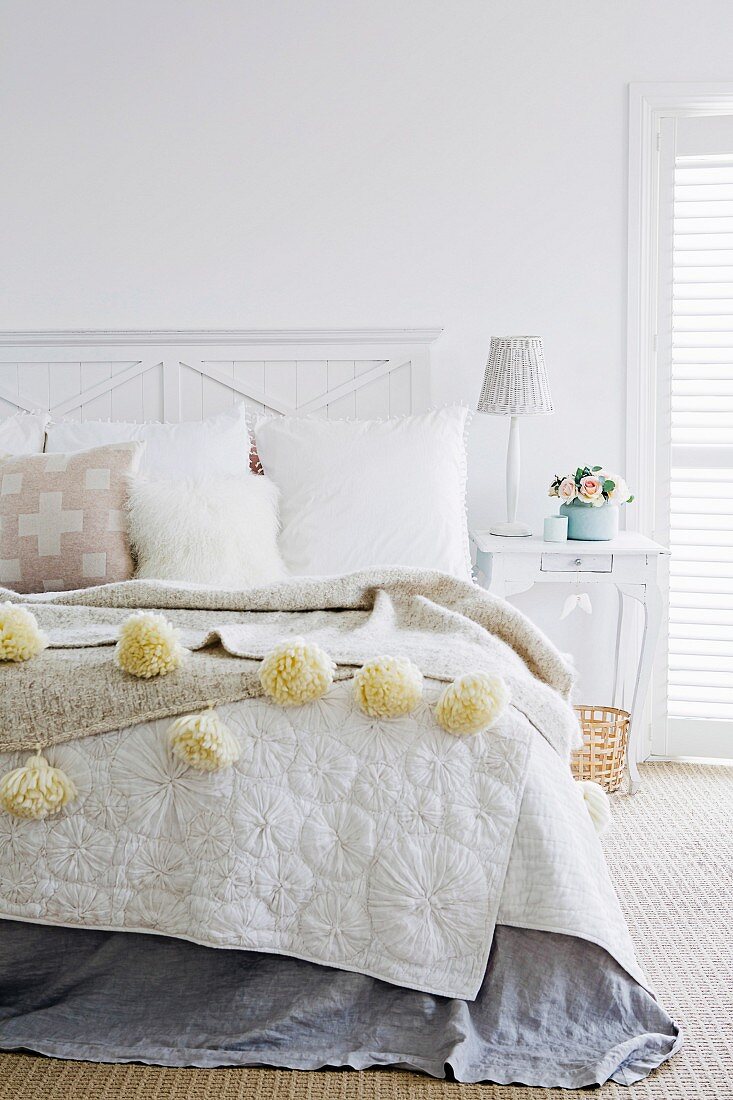 Fur cushion and blanket with woollen pompoms on bed in light-flooded bedroom
