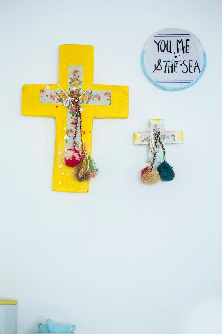 Crosses decorated with wooden pompoms and motto on ceramic disc arranged on wall