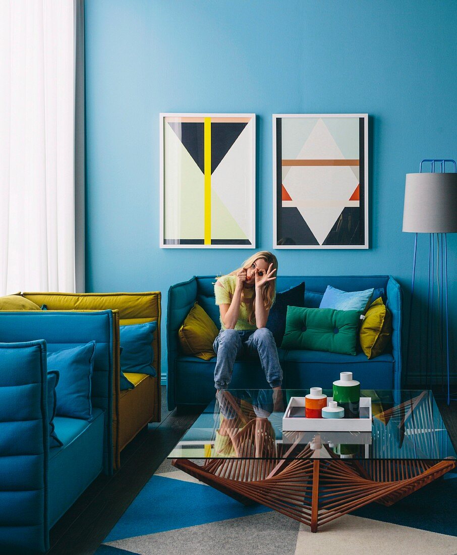 Frau auf blauem Sofa hinter Glastisch und gerahmte Bilder mit geometrischen Mustern an blauer Wand