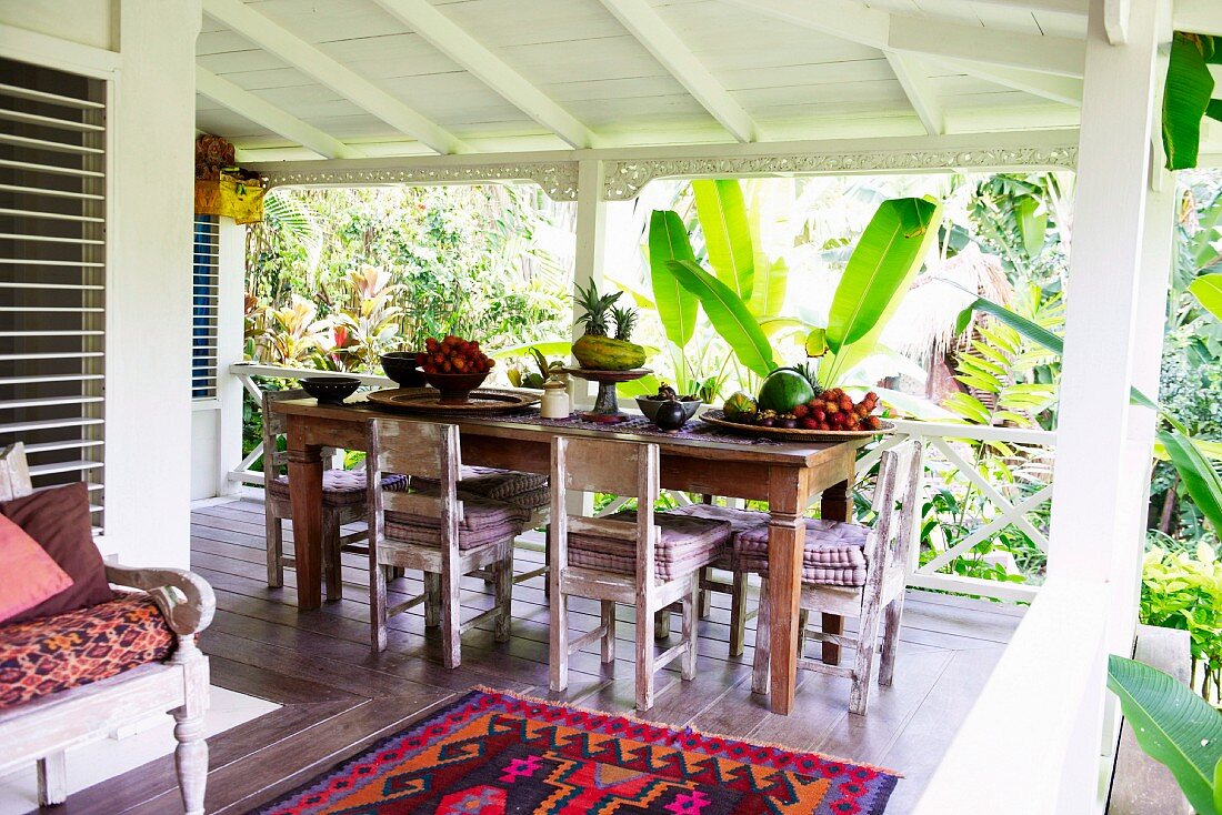 Esstisch mit einfachen Holzstühlen und Obstschalen auf Holzterrasse in tropischer Umgebung