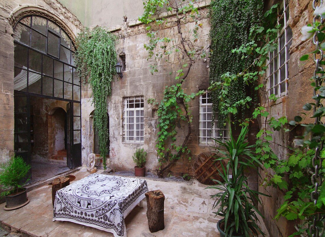 Large table with tablecloth in courtyard of old house with climber-covered walls and glass and steel door in arched doorway