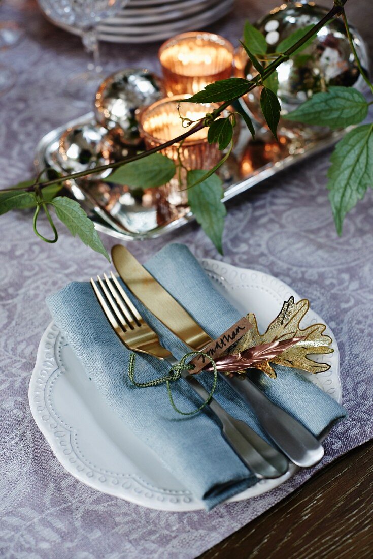 Weihnachtliches Gedeck mit Namensschild auf floral gemusterter Tischdecke und Grünpflanzendeko im Vordergrund