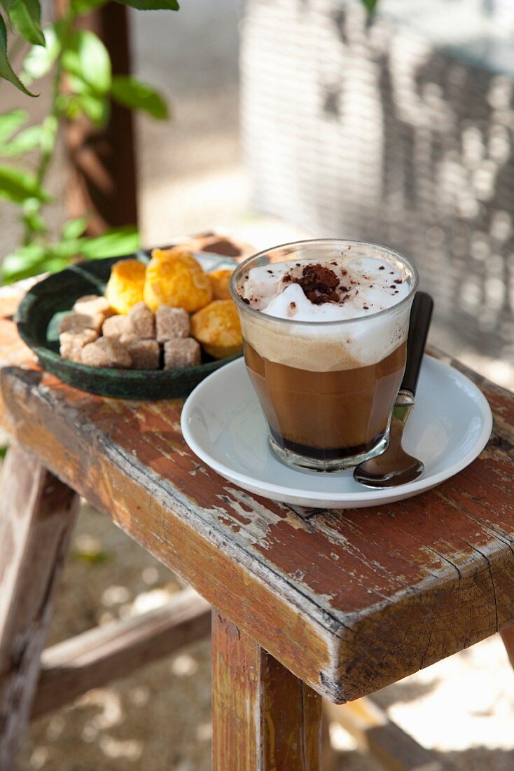 Cappuccino im Glas und Konfekt in Schale auf rustikalem Holzschemel