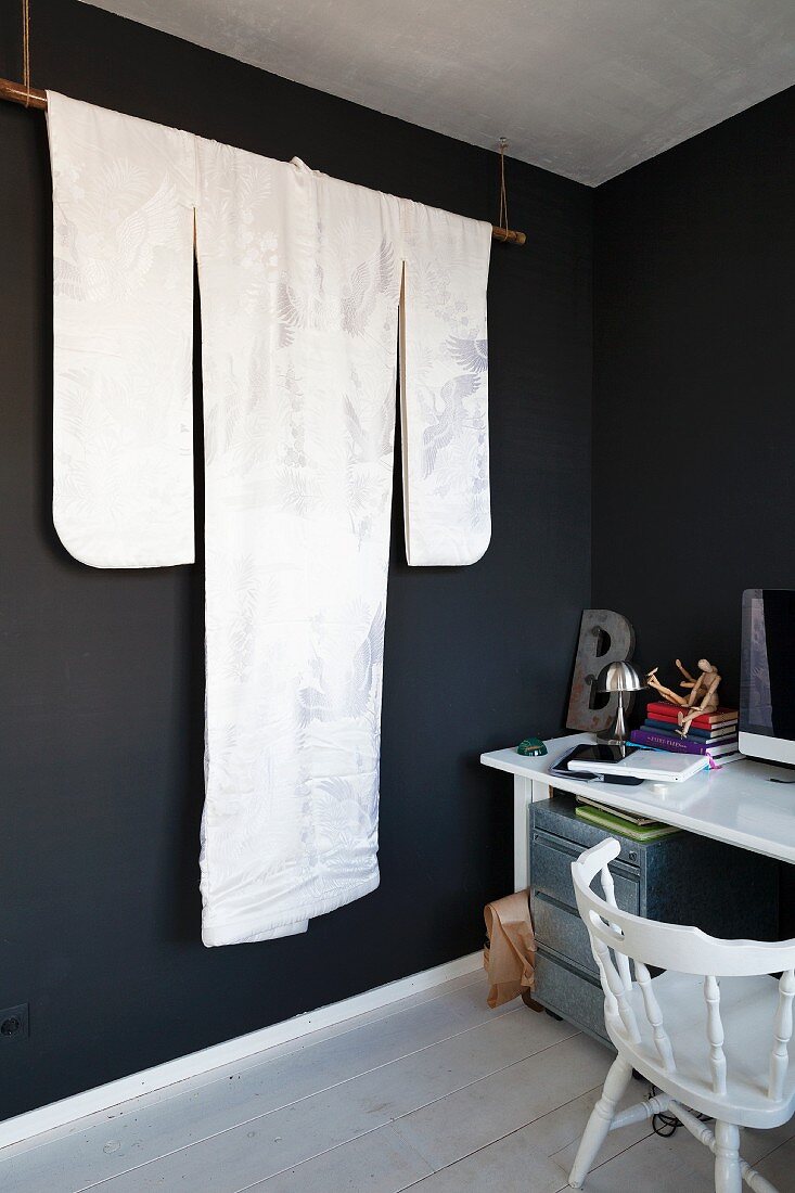 Von Decke abgehängte Stange mit edlem Kimono in Weiß vor schwarz getönter Wand, seitlich schlichter Arbeitsplatz