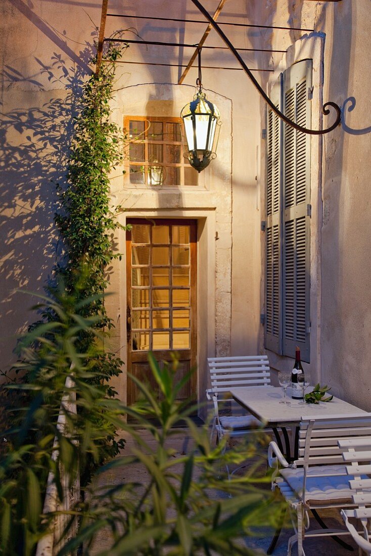 Lantern-style lamp above table and chairs against exterior house wall with closed window shutters