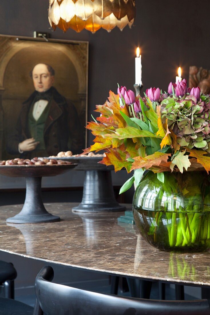 Spherical vase of flowers and dark grey ceramic dishes on stone table top in front of old portrait