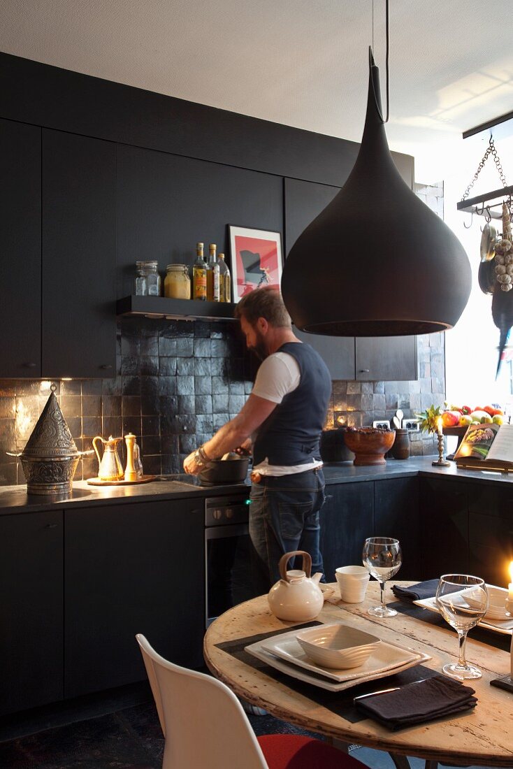 Gedeckter Tisch unter schwarzer Designer-Pendelleuchte und kochender Mann vor schwarzer Einbauküche
