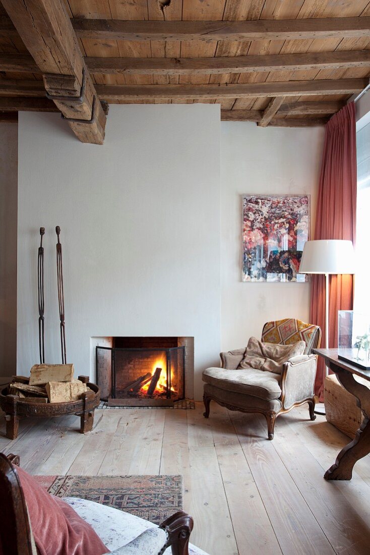 Comfortable antique armchair next to open fire in rustic interior with wooden floor and wood-beamed ceiling