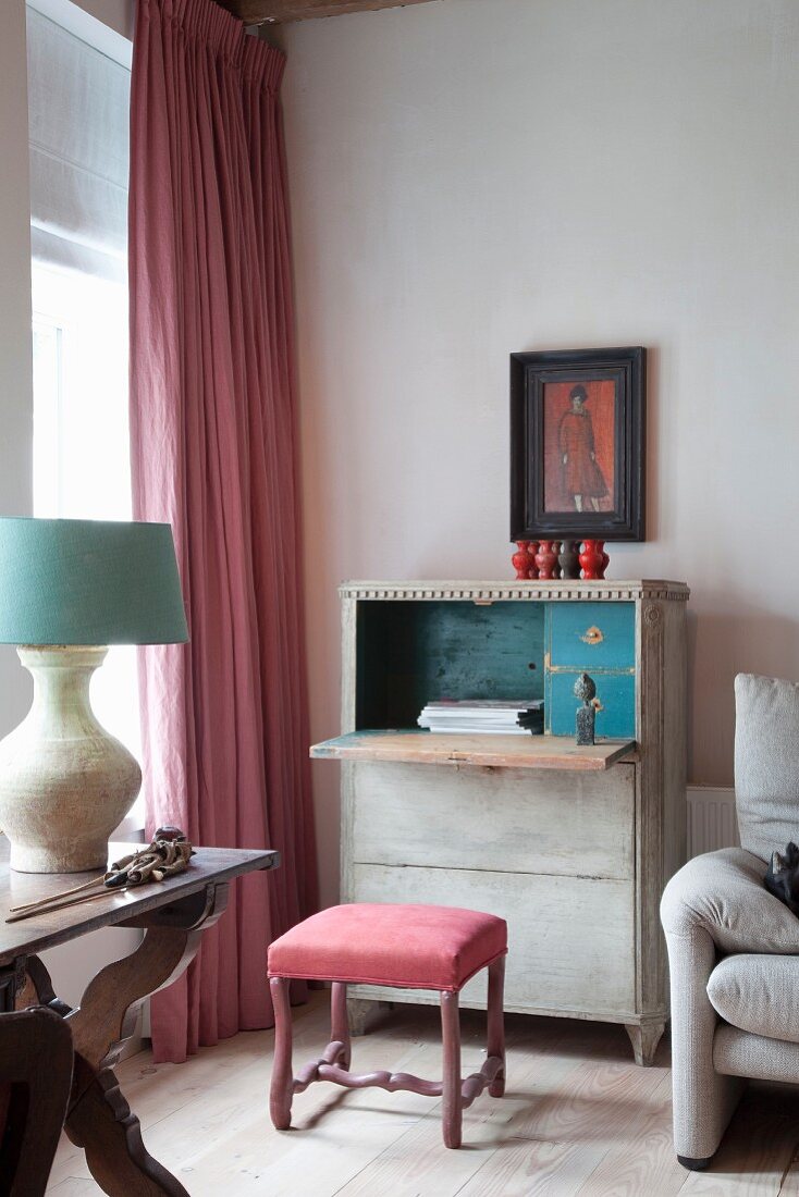 Antique upholstered stool at vintage writing desk next to floor-length curtain