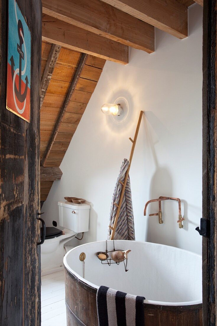 View through open door of vintage bathtub in rustic attic room with wood-beamed ceiling
