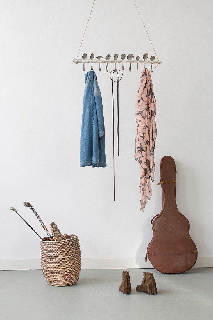 Curved vintage spoons inserted through suspended wooden rod used as coat rack above old guitar case and hockey sticks in basket
