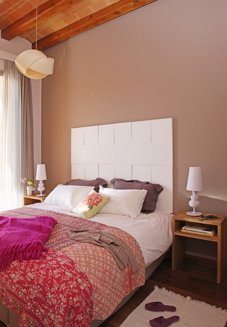 Patterned, patchwork-style cover on double bed with white panel headboard against pastel brown bedroom wall