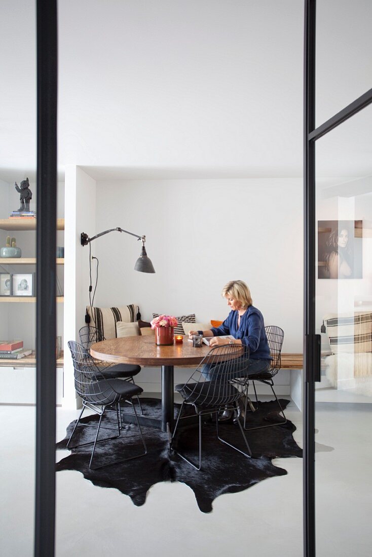 Blick durch offene Glastür auf Frau an rundem Tisch, moderne, schwarze Stühle aus Metallgittern auf Tierfellteppich