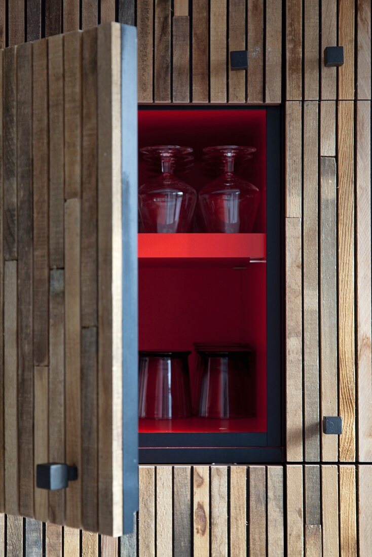 Open kitchen cupboard with wooden door, red-painted interior and glasses on shelves