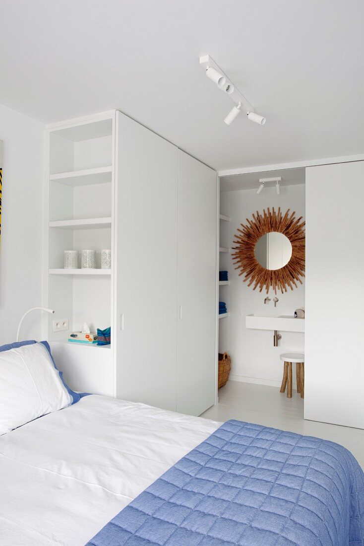 Blue bedspread on double bed, custom wardrobe with shelving element and small bathroom behind open sliding door