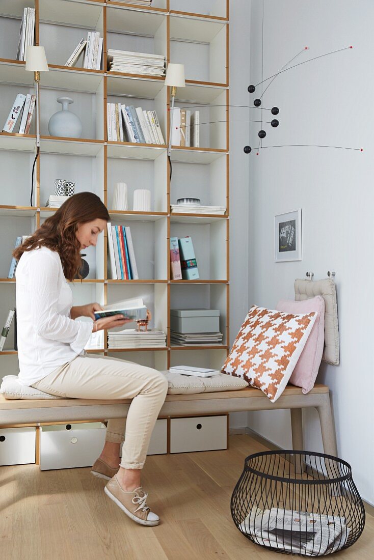 A woman reading on an upholstered bench with a cushion hanging on the wall from hooks in front of a light painted designer shelf system