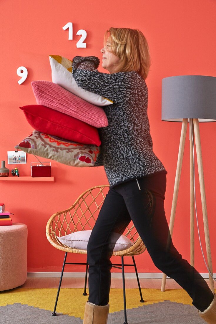 A woman walking past a coral-coloured wall with a stack of cushions with a simple floor lamp and a retro-style wicker chair in the background