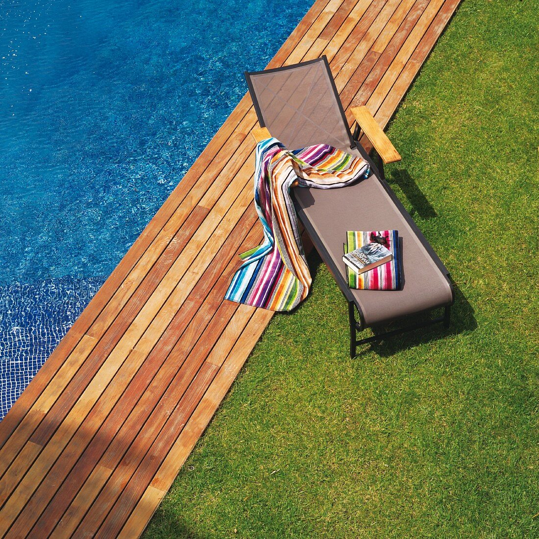 Rasen und Holzdeck an Poolrand mit bunt gestreiften Badetücher auf moderner Sonnenliege