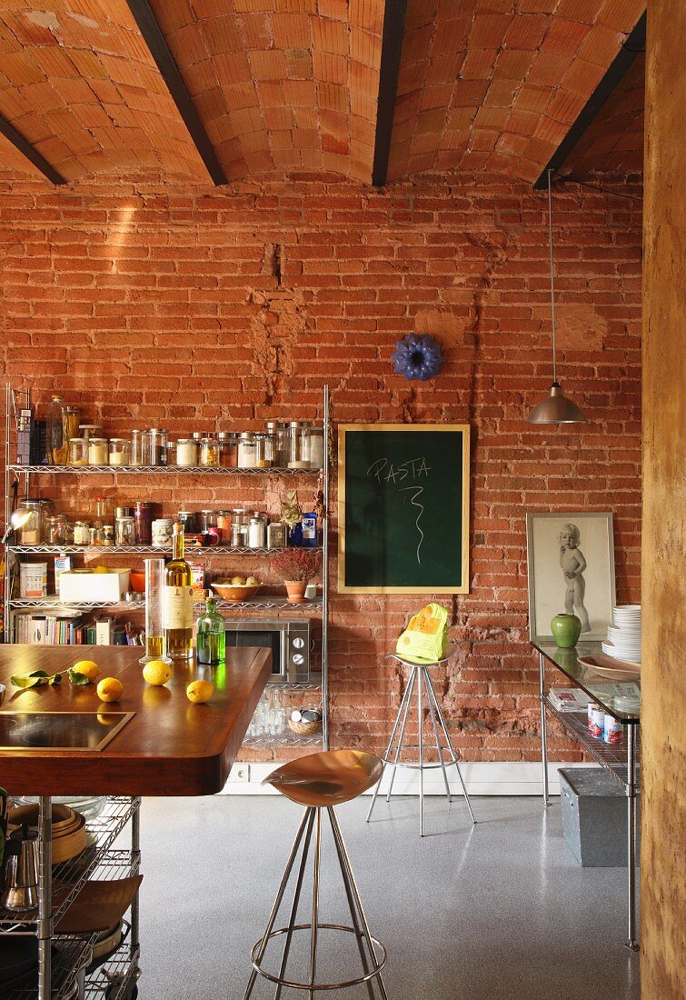 Barhocker mit Kupfersitz an Frühstückstheke in einer Loftküche; offenes Metallregal und Tafel vor Backsteinwand