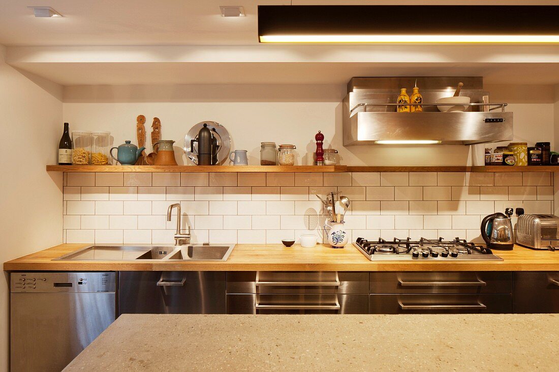 Blick über Theke auf Küchenzeile mit Holzarbeitsplatte vor weiss gefliester Spritzschutzwand, darüber Wandbord aus Holz und Dunstabzug