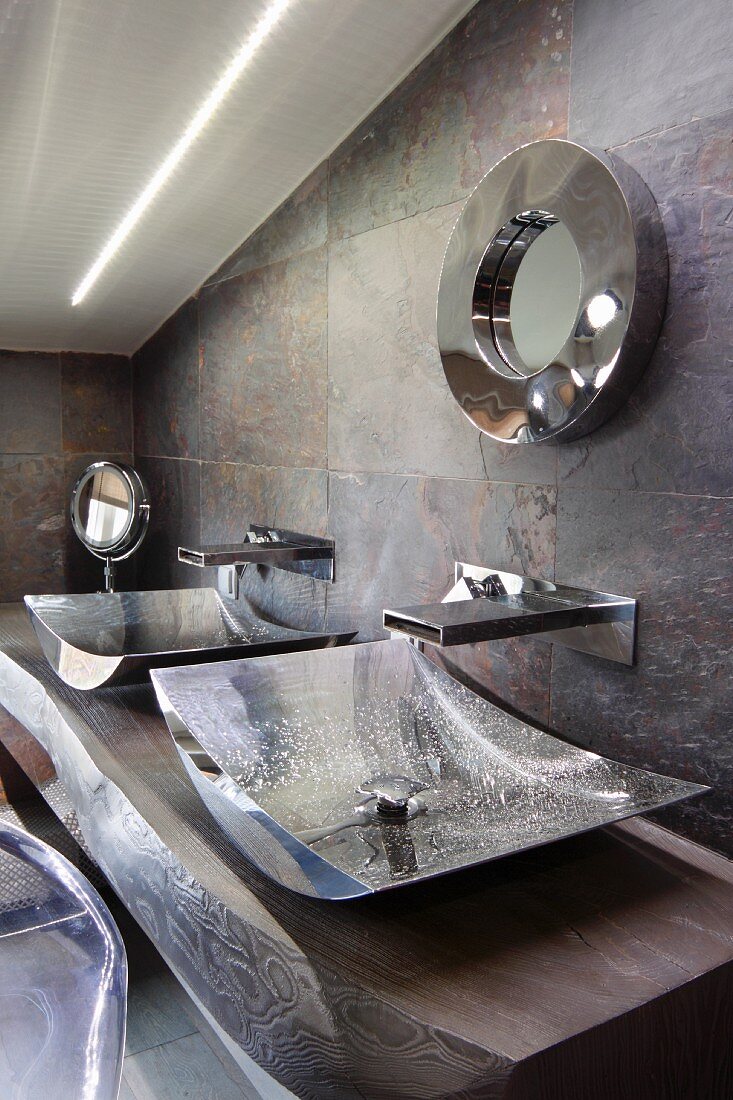 Two curved chrome basins on solid wooden washstand against stone-tiled wall